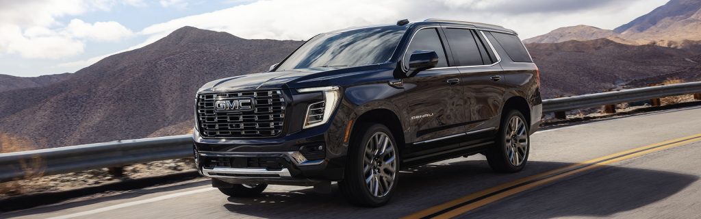 Black GMC Yukon Denali SUV driving on a scenic mountain road, highlighting its luxurious design and bold exterior features.