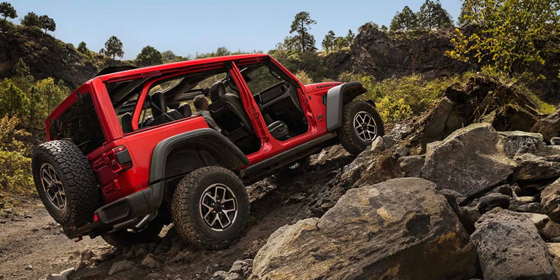 Red Jeep Wrangler SUV climbing over large rocky terrain on an off-road trail, with open doors and rugged outdoor landscape in the background.