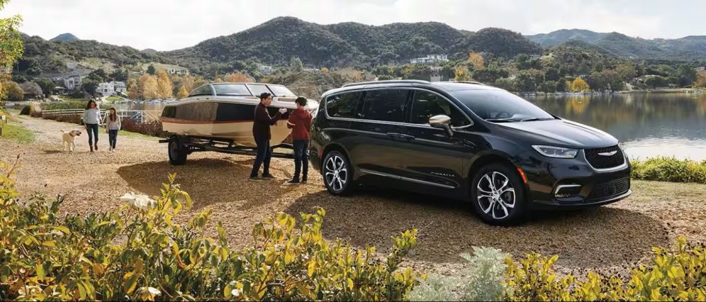 Chrysler minivan towing a boat near a lakeside mountain landscape.